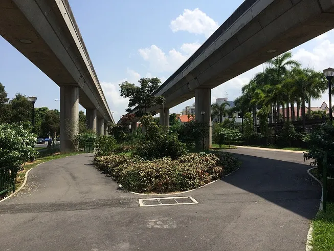 An underpass with two concrete railway tracks overhead is surrounded by lush greenery and trees.
